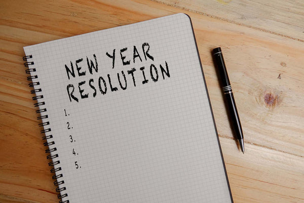 A pen and notepad sit on a wooden table with the words, “New Year Resolution” written on it with five blank lines