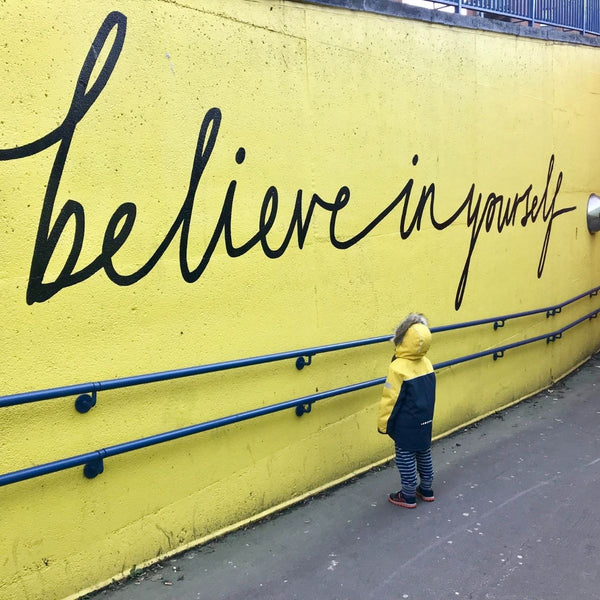 A boy in a coat staring at a yellow wall with the phrase, “Believe in yourself” painted on it